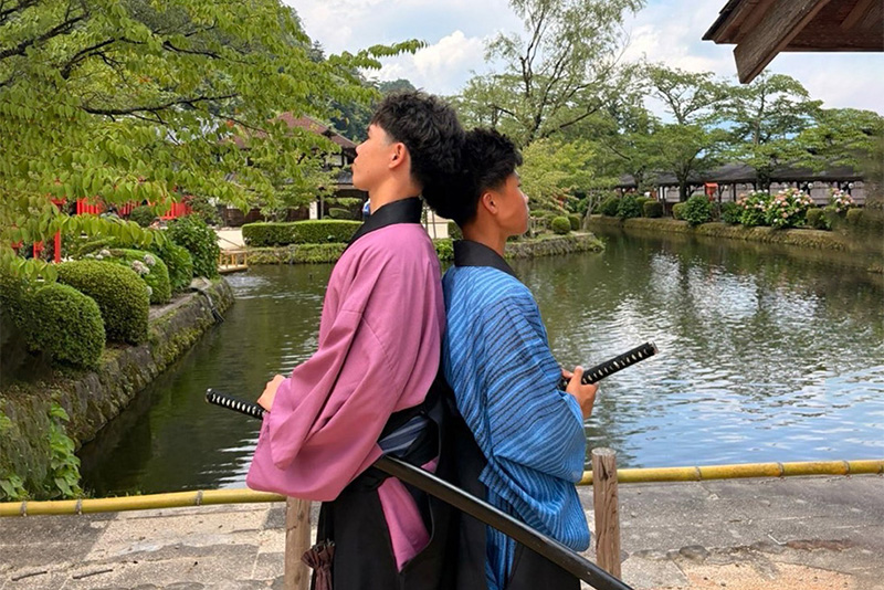 Two students wearing traditional Japanese clothing and posing with swords by a pond.